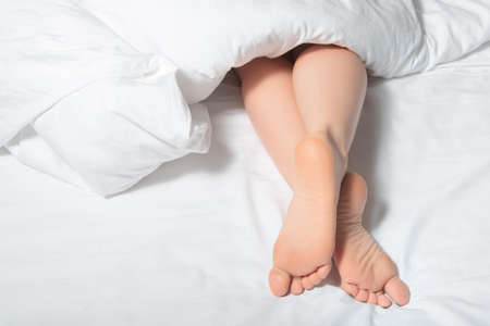 woman's feet under the white blanket. woman's leg looks out from under the covers on a white bed....