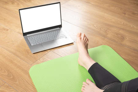 woman sits on yoga mat with her legs stretched out and looks into mockup of laptop with blank white screen. woman with laptop computer exercising at living roomの写真素材