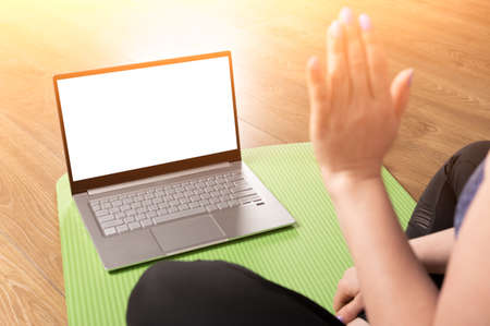 Woman greeting fitness coach online on laptop blank white screen before online yoga lesson. Woman Training At Home Online Via Laptop, Waving Hand At Camera, Greeting Fitness Instructorの写真素材