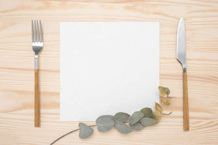 Knife and fork with white paper for menu or recipe text and gold eucalyptus branch on wood table. Mockup Blank card and cutlery on rustic wooden table backgroundの写真素材