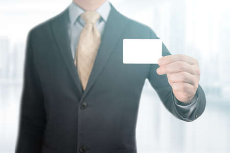 Hand holding blank business card. Handsome businessman in black suit showing his mockup credit card to make a payment. man holding showing blank business card or name card. blurred office backgroundの写真素材