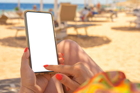 Closeup of young woman's hands using white mobile smartphone Mockup on the beach, beach resort and Sea in the background. woman holding mobile phone with blank desktop screen while sitting on beachの写真素材