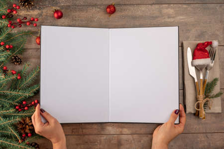 Woman hands holds christmas menu mockup. Christmas Recipe Menu mockup on rustic wooden background. Christmas menu concept with cutlery decorated with tree twigs and baublesの写真素材