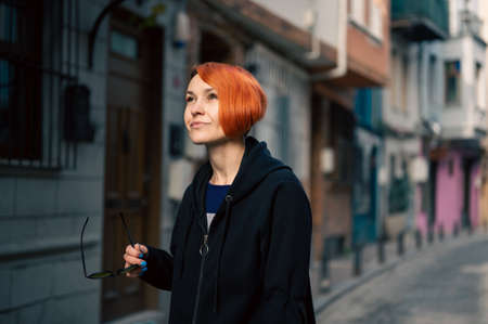 Young lesbian woman confidently walks down street and looks ahead. Young cheerful woman with bright red hair walks down narrow street. attractive woman walks along narrow city streetの写真素材