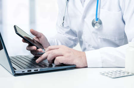Male doctor with laptop and cellphone at desk in medical office. Physician using laptop at desk in...