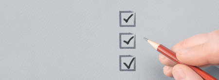 hand with pencil writing check mark on Checklist. Hand with pencil and check boxes on gray paper background. Male hand choosing three of three options.の写真素材