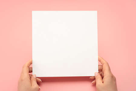 Overhead shot of woman hands holding square blank paper sheet on pink table. Woman hands with mockup...