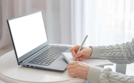 Side view and close up of woman's hands using laptop with blank white screen and writing in notepad....