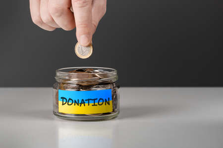 Donation for Ukraine concept. Help and financial assistance to Ukraine. donation jar with Ukrainian flag. Ukrainian war, donations, Hand putting Coins in glass jar with donationの写真素材