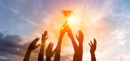 Success of teamwork, joint achievement of goal in business and life. Winning team is holding trophy in hands. Silhouettes of many hands in sunset.の写真素材