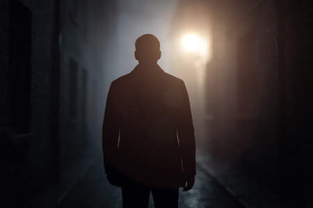 Lonely man in shadowy alley at night, facing glowing streetlight, showing concept of urban insecurity, violence, crime, risk, thriller sceneの写真素材