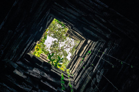 Low angle view looking up inside ancient stone tower square opening sky green trees sunlight perspective architecture ruins mystic atmosphere geometric frameの写真素材
