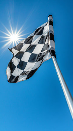A vibrant checkered flag flutters proudly in the breeze against a backdrop of a clear blue sky, illuminated by the bright sun, signaling the excitement of racing.の素材