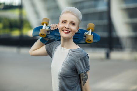 Young woman with short blonde hair smiling on the fashion background and holding little penny skateboard behind her head. The girl in joyful feelings. Outdoors, lifestyle. The city urban outdoor backgroundの写真素材
