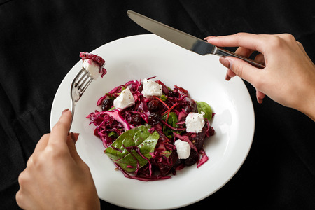 Top view salad with beet cabbage and mozzarella. Woman hand holding folk. White plate with fresh beet salad with beet cabbage and mozzarella on black table. Restaurant food. Healthy nutrition.の写真素材