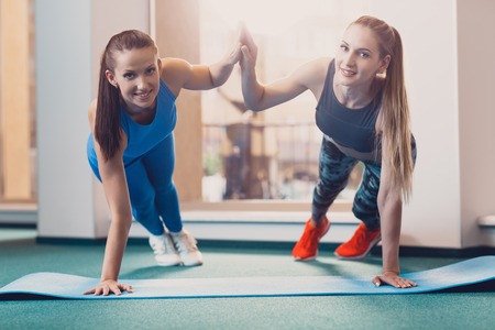 Two happy girls perform sports exercise at workout. Girls greet each other while standing on one hand. Fitness training to improve physical fitness. Sports equipment for training in the gym.の写真素材