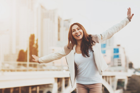 Smiling girl running with hands in air outdoors. A young girl happily runs waving her arms to the side in the fresh air in the park. Happy girl stands against the background of houses and the bridge.の写真素材