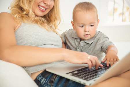 Happy young mother playing with her son the couch. Mom and son are watching a baby video on a laptop. Young family resting on the sofa with laptop. Happy family spends their holiday lying on the couchの写真素材