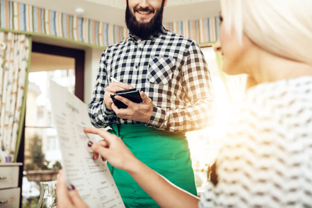 Close-up of a waiter accepting an order from girl. Man taking order from girl in cafe. Waiter serving concept. Young beautiful woman thinking of food to order in front of waiter holding tablet.の写真素材