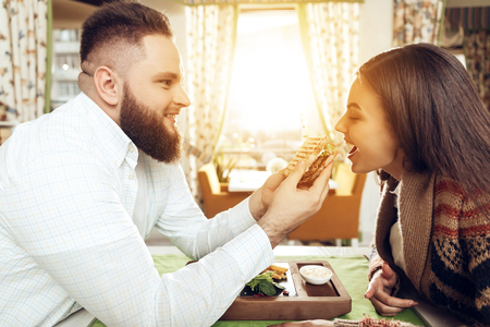 Happy man and woman are having lunch in a restaurant. Smiling man and woman couple enjoying their romantic lunch. Guy feeds the girl with a sandwich. The concept of the restaurant business.の写真素材