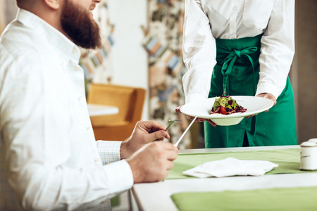 Girl waiter serves his dish in restaurant to a man. The waiter serves in the restaurant of a smiling bearded man. Service serving meals to the visitor in the restaurant.の写真素材