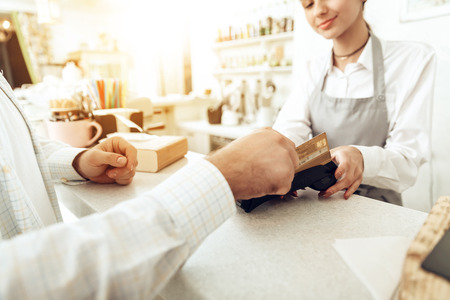 Cheerful cashier using digital device for payment. A smiling girl waiter calculates a man in a cafe with a bank card. The concept of the restaurant business and service maintenanceの写真素材