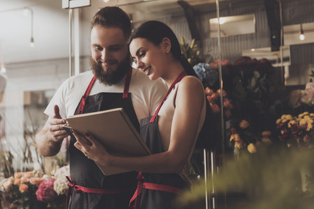 Portrait of smiling florists man and woman. Beautiful florist girl makes working notes in notebook. Young bearded male florist helps colleagues in work flower shop holding a mobile phone in his hand.の写真素材
