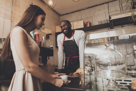 Smiling barista man gives a girl her order. Afro-American barista male is giving a young girl her croissant for coffee. The concept of restaurant business and quality of serviceの写真素材