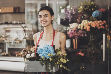 Portrait of beautiful girl florist in flower shop. Young smiling girl florist collects flower basket for a customer in a flower shop. Work that brings pleasure. Flower business concept.の写真素材