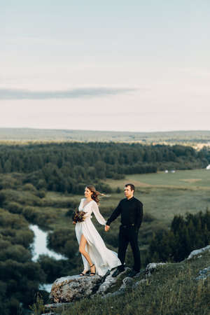 A beautiful fairy-tale happy bride with a bouquet and a wreath and a stylish groom on the background of rocks in the mountains. Boho wedding in nature.の写真素材