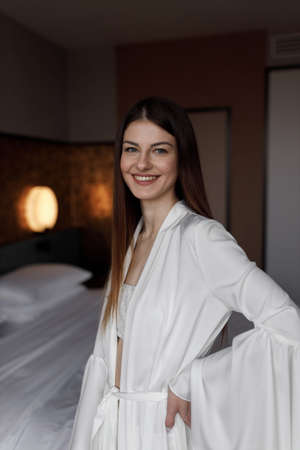 A young girl poses and smiles before the wedding. Beautiful morning of the bride in a light boudoir dress in a hotel on the bed.の写真素材