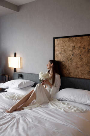Beautiful morning of the bride in a light boudoir dress in a hotel on the bed. A young girl poses and smiles before the wedding.の写真素材