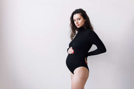 A young beautiful pregnant woman in a black bodysuit, on a white wall. The concept of a fashionable image for pregnant women.の写真素材