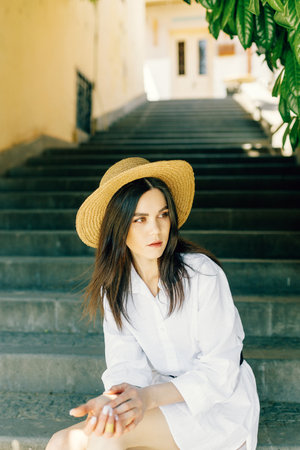 a cute girl in a hat is sitting on the stairs of the old town. Brunette in a white dress walks the streetの写真素材