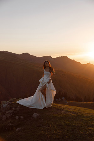A woman elegantly attired in a stunning wedding dress is standing atop a picturesque hill as the sun sets beautifully on the horizonの写真素材