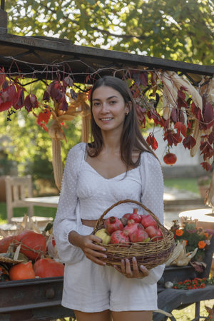 A joyful woman smiles holding a basket of fresh apples, surrounded by autumn decorations at a bustling marketの写真素材