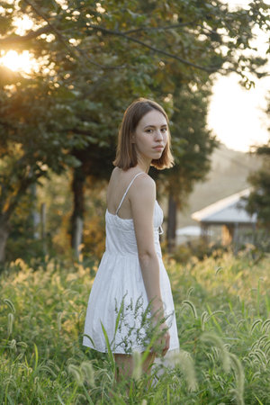 A tranquil and serene portrait of a lovely girl wearing a flowing white dress, embraced by nature during sunsetの写真素材