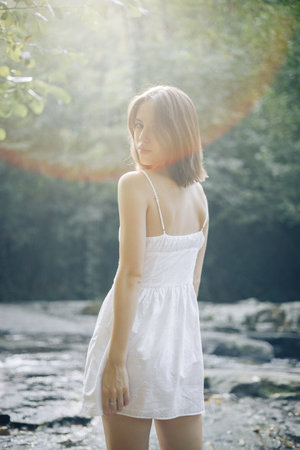 A young woman in a white dress stands by a serene water body, capturing a tranquil moment in nature.の写真素材