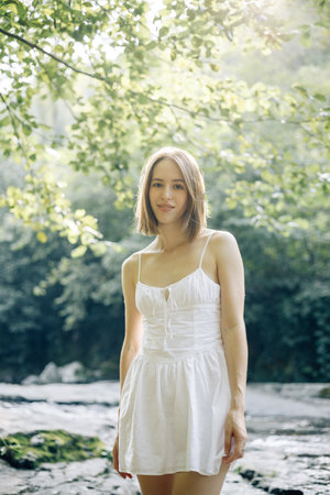 A young woman in a white dress stands by a serene water body, capturing a tranquil moment in nature.の写真素材