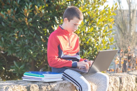 Serious teenage boy with laptop and textbooks doing homework and preparing for the exam in the park among the trees. The concept of heavy learning, a lot of homeworkの写真素材