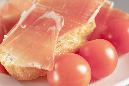 Dry Spanish ham, jamon serrano, Bello, Italian ham prosciutto crudo or parma, chopped layers with bread and cherry tomatoes on a white heart-shaped plate, macroの写真素材
