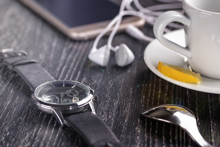Wrist watch and mobile phone with headphones and a cup of tea with lemon on a dark wooden table. The...