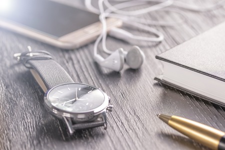 Old dark office desktop with elements for business and work. Wrist watch, mobile phone with headphones, notebook and penの写真素材