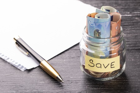 Euro bills and coins in a glass jar with a plate with a blank sheet of paper and a pen on a black wooden table with a sunny reflection on a glossy surface. Savings conceptの写真素材