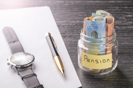 Glass jar with coins and euro banknotes with the words PENSION. Wrist watch, pen and blank paper. Concept of financial savings for wealthy old ageの写真素材