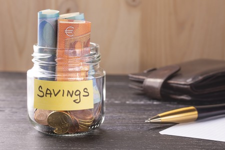 Glass jar with coins and euro notes with the inscription SAVINGS. On the old black wooden table....