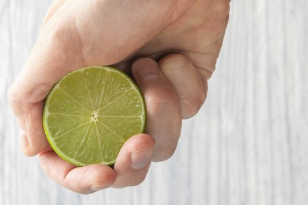 The hand of a man squeezes the juice from a slice of lime half. Juicy drop hanging down. Light wooden background.の写真素材