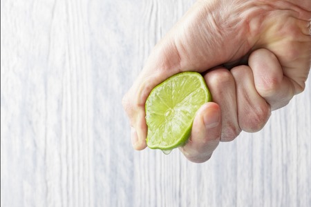 The hand of a man squeezes the juice from a slice of lime half. Juicy drop hanging down. Light wooden background.の写真素材