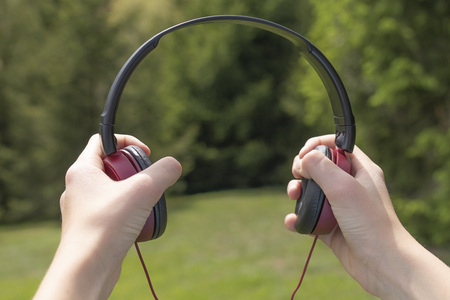 Red-black headphones in the hands against the background of coniferous trees on the green grass and mountains in the distance. Relaxation concept to the sounds of natureの写真素材