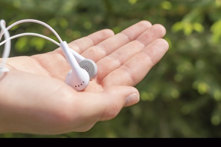 White headphones in the hand of a teenager on the background of coniferous trees in the park. The...